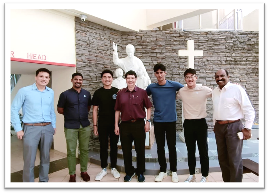 Seven men stand together smiling in front of a stone wall with a white statue and a cross.